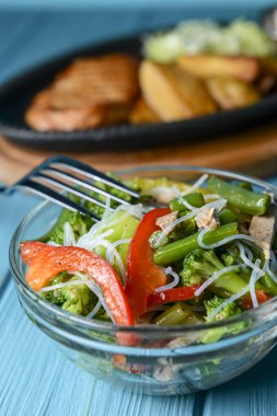 Brokolili sotelenmiş sebze salatası, kuşkonmaz, tatlı biber ve selofan erişte veya mavi arka planda cam bir kasede fensi eriştesi. Akşam yemeği fikri, kızarmış tavuklu biftek ve kızarmış patates..