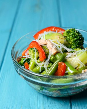 Brokolili sotelenmiş sebze salatası, kuşkonmaz, tatlı biber ve selofan erişte veya mavi arka planda cam bir kasede fensi eriştesi. Akşam yemeği fikri, uzay afişini kopyala.