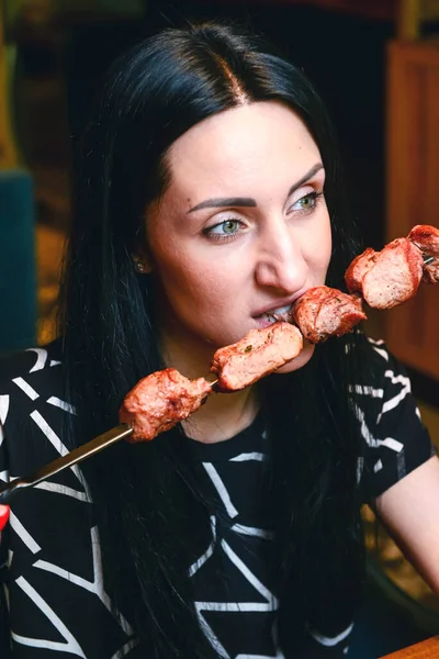 Young woman enjoys grilled meat in a restaurant. Eating shashlik inside, Georgian cuisine concept. Brunette caucasian girl, eat out alone concept, social distancing.