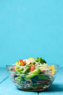 Brokolili sotelenmiş sebze salatası, kuşkonmaz, tatlı biber ve selofan erişte veya mavi arka planda cam bir kasede fensi eriştesi. Akşam yemeği fikri, uzay afişini kopyala.