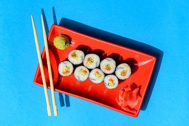 Japon suşi ruloları kırmızı bir tabakta turşulu zencefil ve wasabi sosuyla servis ediliyor. Renkli mavi arka planda. Japon mutfağı konsepti. Geleneksel suşi maki rulo tarifi.