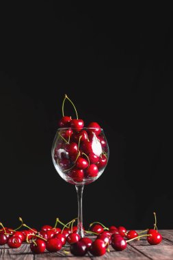 Siyah arka plan üzerinde, uzun bir şarap kadehinde, ahşap bir masada mevsimlik taze kiraz meyvesi. Uzay tasarım afişini kopyala. Yaz konsepti, kırsal tarzda taze kırmızı kiraz..