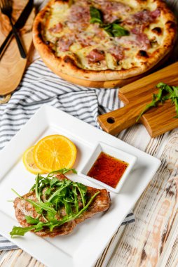 Taze yeşil roka, portakal dilimleri, domates sosu ve jambonlu taze pizza ile servis edilmiş ızgara biftek aromalı gurme. Beyaz bir tabakta. İtalyan mutfağı kavramı.