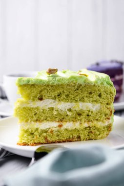 Fıstıklı pasta, beyaz tabakta fındıklı kremalı pasta. Fıstıklı tatlı pasta. Ahşap zemin üzerinde yeşil tatlı. Arka planda bir fincan kahve ve lavantalı kek..