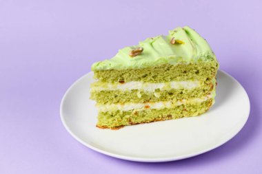 Fıstıklı pasta, beyaz tabakta fındıklı kremalı pasta. Fıstıklı tatlı pasta. Pastel mor arka planda yeşil tatlı. Arka planda bir fincan kahve ve lavantalı kek..