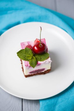 Üzerine krema ve taze kiraz konmuş, beyaz bir tabakta servis edilen, beyaz ahşap arka planda duran, kirazlı gül rengi cheesecake. Lezzetli tatlı zamanı, pastayı kapat, uzay afişini kopyala.