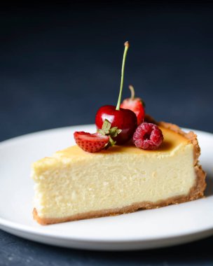 Lezzetli bir parça New York usulü vanilyalı cheesecake. Üstünde böğürtlen var. Beyaz tabakta servis edilmiş, seçici bir odak noktası. Geleneksel New York Cheesecake tarifi. Siyah arkaplanda.