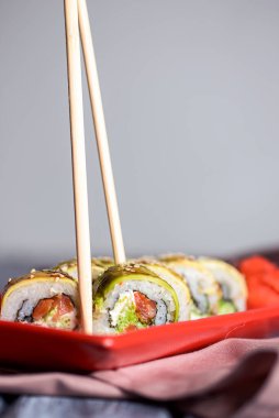 Uramaki suşi rulosu füme yılan balığı, unagi, avokado ve susam tohumlarıyla kaplı yeşil ejderha açık ahşap zemin üzerinde kırmızı bir tabakta servis edilir. Japon mutfağı kavramı.