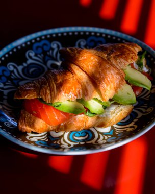 Kırmızı masanın üzerinde renkli bir tabakta servis edilen somon ve avodaco ile kruvasan. Fransız mutfağı, öğle yemeği fikri..