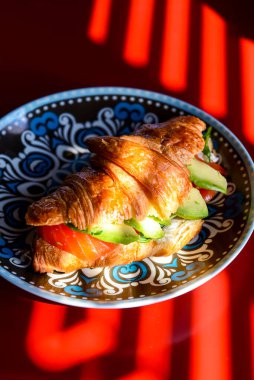 Kırmızı masanın üzerinde renkli bir tabakta servis edilen somon ve avodaco ile kruvasan. Fransız mutfağı, öğle yemeği fikri..