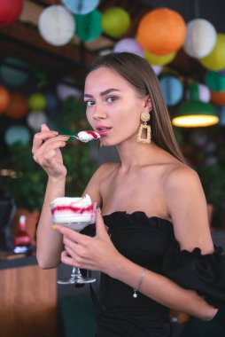Lokantadaki güzel, seksi esmer kadın ya da dondurmalı, tiramisu tatlısı olan kafe. Siyah elbiseli, makyajlı, tatlı yiyen genç bir kadının moda fotoğrafı. Dışarıda yemek konsepti.