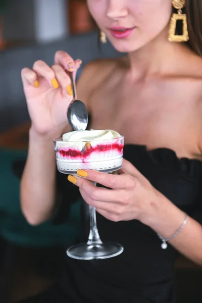 Beautiful sexy brunette woman in restaurant or cafe with ice cream tiramisu dessert. Fashion photo of young woman in black dress with hairstyle and makeup, eating dessert. Eat out concept.