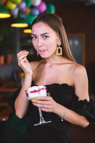 Beautiful sexy brunette woman in restaurant or cafe with ice cream tiramisu dessert. Fashion photo of young woman in black dress with hairstyle and makeup, eating dessert. Eat out concept.