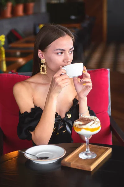 Beautiful sexy brunette woman in restaurant or cafe with ice cream tiramisu dessert and a cup of . Fashion photo of young woman in black dress with hairstyle and makeup eat out concept.