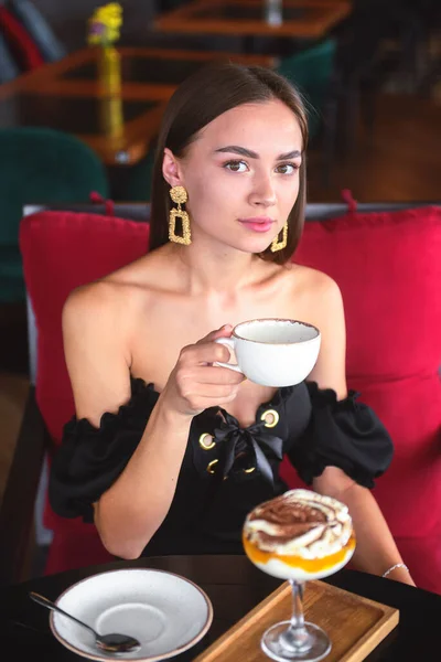 Beautiful sexy brunette woman in restaurant or cafe with ice cream tiramisu dessert and a cup of . Fashion photo of young woman in black dress with hairstyle and makeup eat out concept.