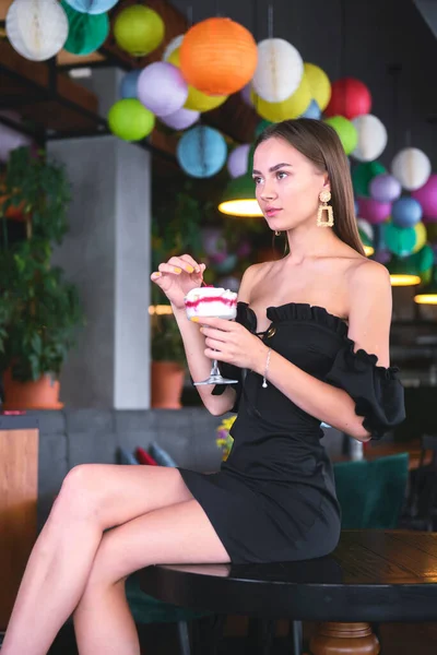 Beautiful sexy brunette woman in restaurant or cafe with ice cream tiramisu dessert. Fashion photo of young woman in black dress with hairstyle and makeup, eating dessert. Eat out concept.