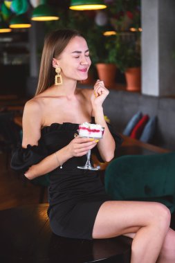 Lokantadaki güzel, seksi esmer kadın ya da dondurmalı, tiramisu tatlısı olan kafe. Siyah elbiseli, makyajlı, tatlı yiyen genç bir kadının moda fotoğrafı. Dışarıda yemek konsepti.