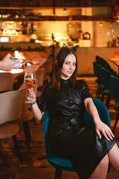 Portrait of young gorgeous woman drinking champagne in glass and looking with smile, enjoying her leisure time inside a restaurant. Girl in a black dress with cat ears.