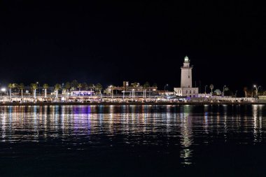 Işıklandırılmış La Farola deniz feneri, palmiye ağaçları ve sakin liman sularına yansıyan tatil ışıkları ile Malaga limanının geniş gece manzarası