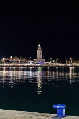 Malaga limanındaki deniz fenerinin (La Farola) gece görüşü, su üzerindeki parıltılı yansımaları, palmiye ağaçlarındaki bayram ışıkları ve derin mavi gökyüzü