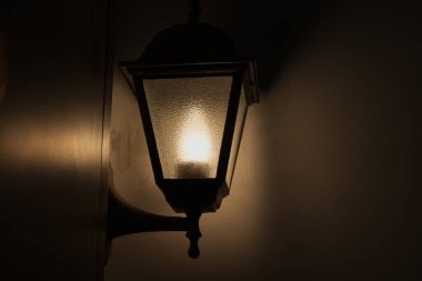 Geleneksel bir siyah taşıma tarzı açık hava lambası koyu bir duvara karşı sıcak bir kehribar ışığıyla parlar. Kalıplı cam pencere yumuşak, davetkar bir atmosfer yaratıyor. Ev güvenliği, konukseverlik ve klasik dekorasyon temaları için mükemmel..