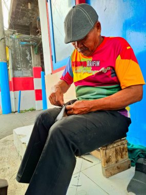 Surabaya, Endonezya, 24 Kasım 2025. Yaşlı bir zanaatkar dikiş dikerek kadın ayakkabılarını onarıyor. Sokaktaki küçük işletmeler.