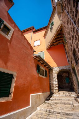 Rovinj, Hırvatistan - 14 Ağustos 2025: Terracotta ve taş duvarlı güneşli bir ara sokak, renkli panjurlar ve Rovinj 'deki dar tarihi evlerden yukarıya doğru uzanan küçük bir kemer yolu.
