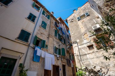 Rovinj, Hırvatistan - 14 Ağustos 2025: Kırık alçı ve yeşil panjurlu eski konut binaları açık mavi gökyüzünün altında yükselirken, çamaşırlar aralarında sallanıyor