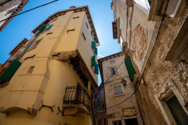 Rovinj, Hırvatistan - 14 Ağustos 2025: Dar dar eski binaların dokulu duvarları, balkonları ve yeşil panjurları dar dar sokaklar arasında açık mavi bir gökyüzüne doğru birleşiyor.