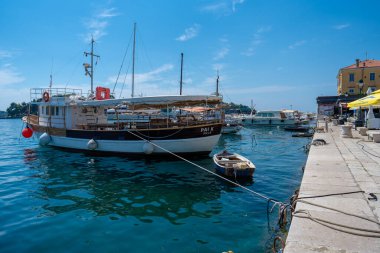 Rovinj, Hırvatistan - 14 Ağustos 2025: Güneşli bir marinaya demirlemiş ahşap balıkçı teknesi, yansıması taş iskele ve diğer rıhtımlı gemilerin yanındaki durgun mavi sularda dalgalanıyor.