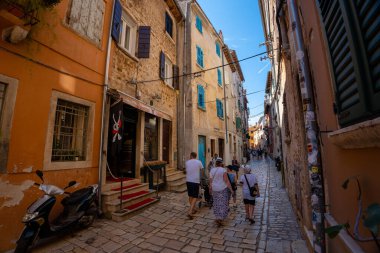 Rovinj, Hırvatistan - 14 Ağustos 2025: Kırsal binalar ve renkli panjurlarla kaplı, turistlerin Korsan Mağarası 'nın önünden geçtikleri ve scooter park ettikleri güneşli bir kaldırım taşı sokak.