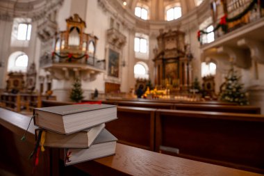 Salzburg, Avusturya - 23 Aralık 2024: Yüksek kemerli pencerelerden süzülen parlak bir barok kilisesi, Noel ağaçları ve güneş ışığı olan ahşap bir sıranın içindeki ilahi kitapları yığını.