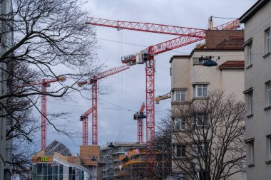 Viyana, Avusturya - 20 Aralık 2024: Bulutlu bir günde gökdelenlerin ve ağaçların üzerinde yükselen kırmızı inşaat vinçleri, bir yerleşim bölgesindeki kentsel gelişimi vurguluyor.
