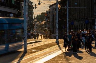 Zagreb, Hırvatistan - 22 Ağustos 2025: Altın gün batımı ışığı canlı bir şehir caddesini yürüyen insanlarla ve hareket halindeki mavi bir tramvayla doldurdu. Ilık ışıltı tramvay raylarını, elektrik direklerini ve eski mimariyi vurgular.,