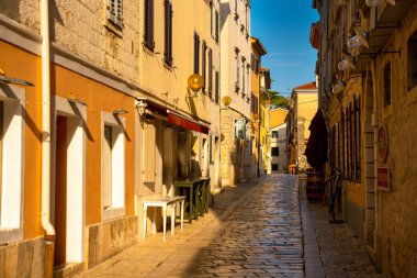 Porec, Croatia - August 13, 2025: A sunlit narrow alley with colorful facades, shutters and stone pavement, glowing warmly in the calm morning light.
