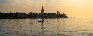 Porec, Hırvatistan - 13 Ağustos 2025: Durgun deniz üzerinde yumuşak bir altın gün batımı yayılıyor, deniz kıyısındaki ufuk çizgisini ve yüksek kilise kulesini serin panoramik bir manzarayla aydınlatıyor.