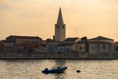 Porec, Hırvatistan - 13 Ağustos 2025: Sıcak akşam ışığı antik taş binalar ve yüksek bir kilise kulesinin üzerine çökerken, küçük bir tekne sakin yansıtıcı su üzerinde sessizce sürükleniyor.