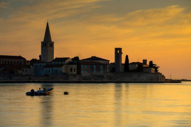 Porec, Hırvatistan - 13 Ağustos 2025: Tarihi bir ufuk çizgisi, kuleler ve ağaçlardan oluşan siluetler ve huzurlu kıyıya yakın sürüklenen küçük bir tekne ile sakin suyun üzerinde altın gün batımı.
