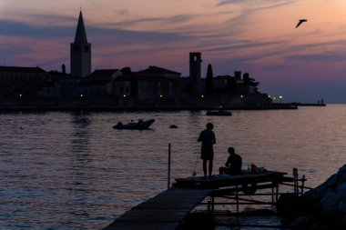 Porec, Hırvatistan - 13 Ağustos 2025: Küçük bir ahşap iskeleden bir baba ve oğulun hafif parlayan kıyı şeridi siluetine karşı balık tuttuğu sakin bir günbatımı sahnesi.