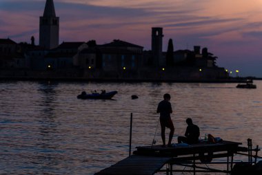 Porec, Hırvatistan - 13 Ağustos 2025: Küçük bir ahşap iskeleden bir baba ve oğulun hafif parlayan kıyı şeridi siluetine karşı balık tuttuğu sakin bir günbatımı sahnesi.