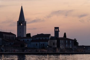 Porec, Hırvatistan - 13 Ağustos 2025: Günbatımında sakin akşam sularına yansıyan yumuşak pastel gökyüzüne karşı tarihi liman binaları ve kilise kuleleri
