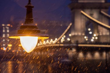 Budapeşte, Macaristan - 6 Ocak 2026: Arkaplanda Zincir Köprüsü 'nün bulanık ışıklarının belirdiği yoğun kar yağışı sırasında gece gökyüzüne parlayan klasik bir sokak lambasının yakın çekim görüntüsü.