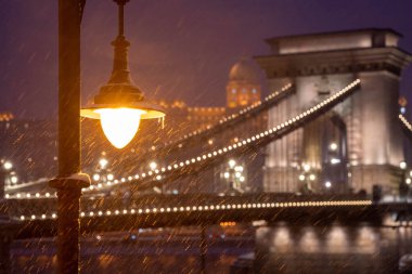 Budapeşte, Macaristan - 6 Ocak 2026: Karlı bir Budapeşte gecesine karşı sıcak bir fener parıldıyor, arka planda ikonik Zincir Köprüsü ve Buda Kalesi şiddetli bir kar fırtınasının içinde parıldıyor.