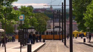 Budapeşte, Macaristan - 24 Mayıs 2025: Güneşli Budapeşte meydanından geçen sarı bir tramvayın manzarası, yayalar, yeşil ağaçlar ve uzak bir binicilik heykeli ile çevrili.