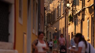 Rovinj, Hırvatistan - 15 Ağustos 2025: Sıcak güneş ışığı dar ve tarihi bir Rovinj caddesini yayalar ve yıpranmış taş cepheler ve geleneksel kepenklerle yürüyerek geçen bir aile olarak yıkıyor.