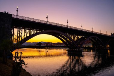 Maribor, Slovenya - 9 Ağustos 2025: Slovenya 'nın Maribor kentindeki Stari Most' un (Eski Köprü) çarpıcı bir günbatımında çekilmiş alçak açılı atmosferik çekimi. Çelik kemerler parlak bir siluet oluşturur, eğimli bir gökyüzüne karşı, mordan altına doğru solan.