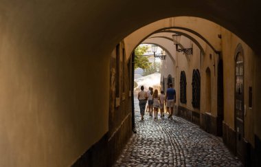 Ljubljana, Slovenya - 10 Ağustos 2025: Bir aile Ljubljana 'da dar, kemerli bir kaldırım geçidinden geçerek, öğleden sonranın sıcak parıltısında güneşli bir sokağa doğru yürüyor.