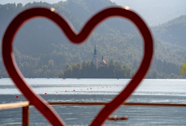 Aşkın sembolünü görkemli bir ortamda yakalayan bu resim, Bled Island 'daki büyük bir kırmızı kalp şekli tarafından çerçevelenmiş ikonik Hac Kilisesi' ni yansıtıyor. Bulanık ön plan, parlak mavi suya ve yeşil Mou 'ya dikkat çekiyor.