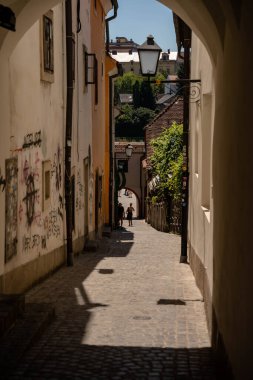 Slovenya 'nın Maribor kentindeki güneşli bir kaldırım taşına gölgeli bir kemerden bakan pitoresk dikey bir manzara. Dar geçitte yıpranmış duvarlar, kentsel grafiti, eski bir demir sokak lambası ve sürünen sarmaşıklar yer alıyor. İki yaya yürüyor.