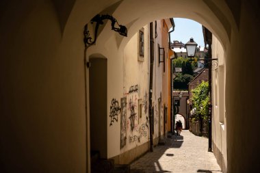 Slovenya 'nın Maribor kentindeki karanlık, kemerli bir geçitten geçen dar, güneşli bir kaldırım taşı caddesini çerçeveleyen yatay bir çekim. Yüksek kontrastlı kompozisyon, taş kaldırımın dokusunu ve kentsel grafiti ile süslenmiş yıpranmış duvarları vurguluyor. Bir...